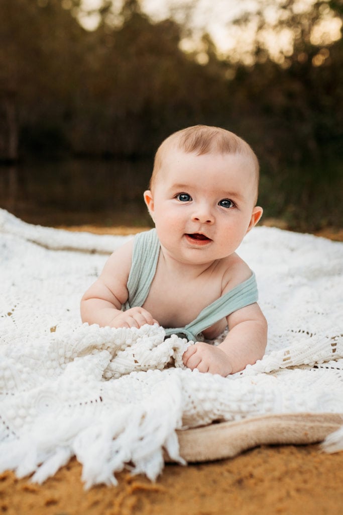 Family Photographer, a baby looks up and smiles while crawling on a blanket outdoors