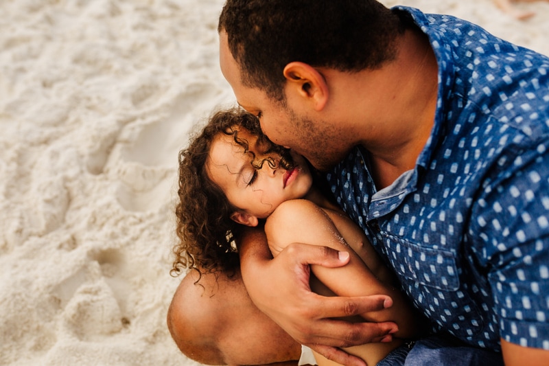 Family Photographer, a dad kisses his young daughter on the cheek as she sleeps in his arms at the beach